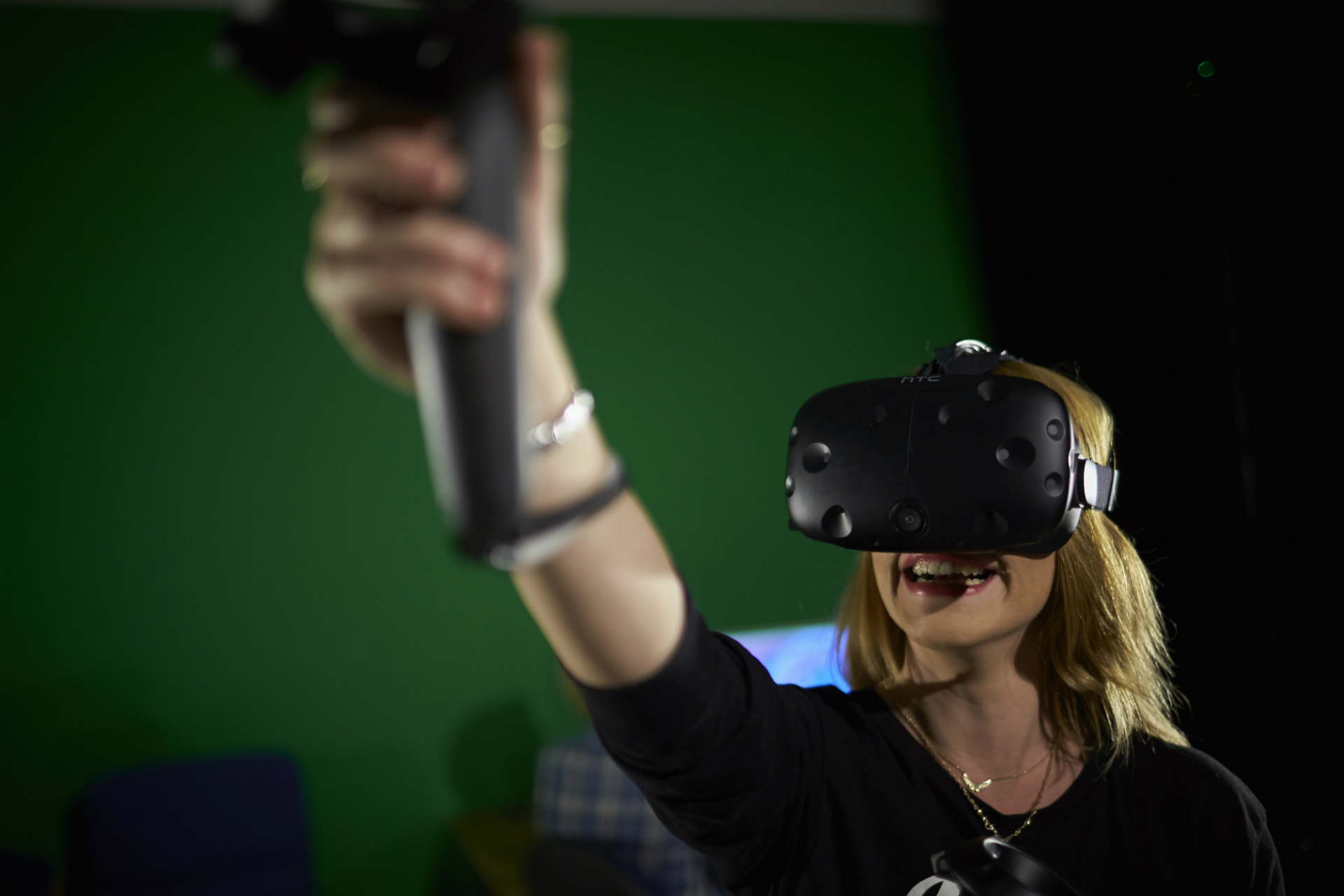A female student, standing in front of a green background, uses a VR head-set. She holds a controller up to the lens and smiles.
