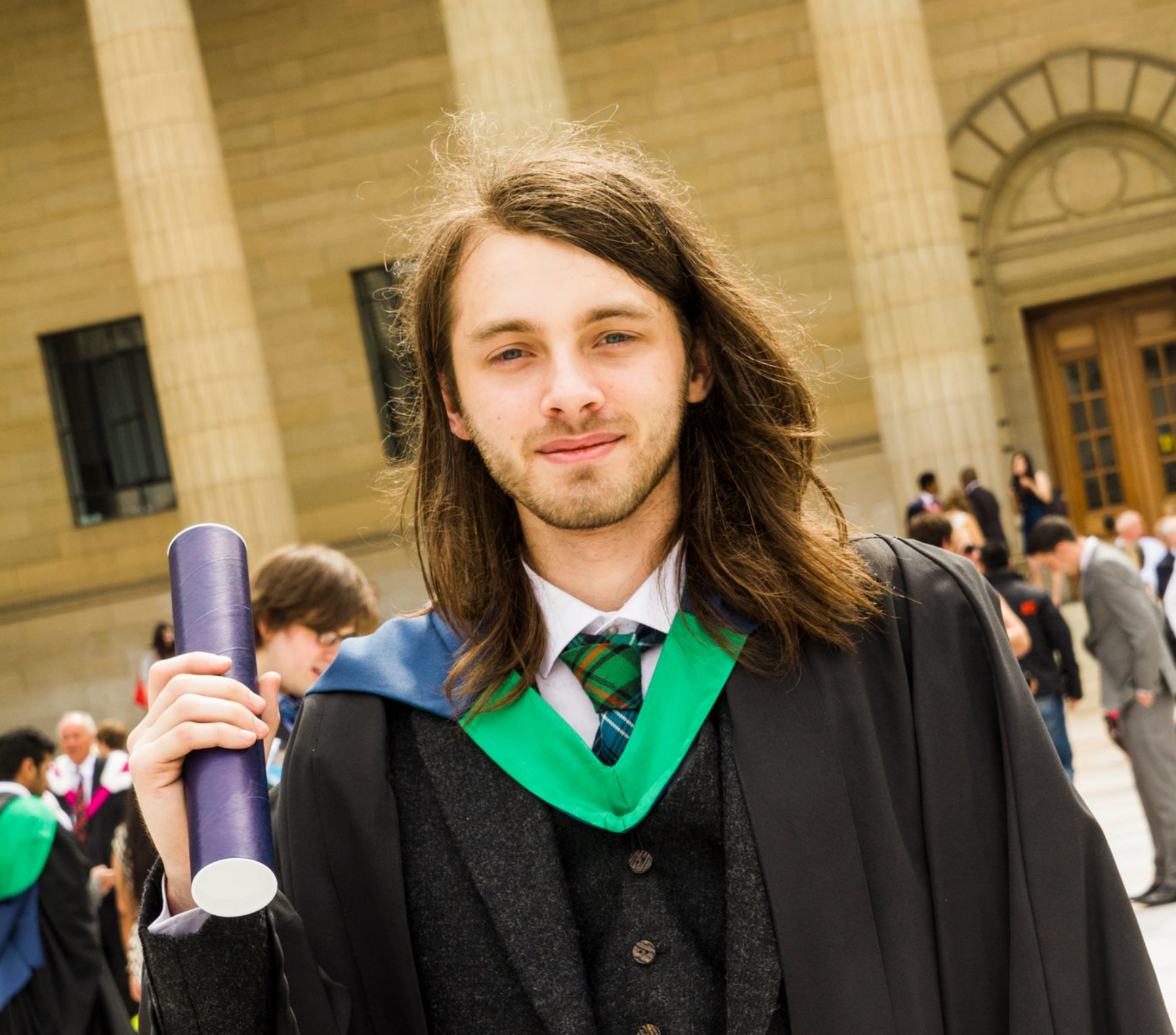 Male wearing graduation robes, holding scroll and smiling