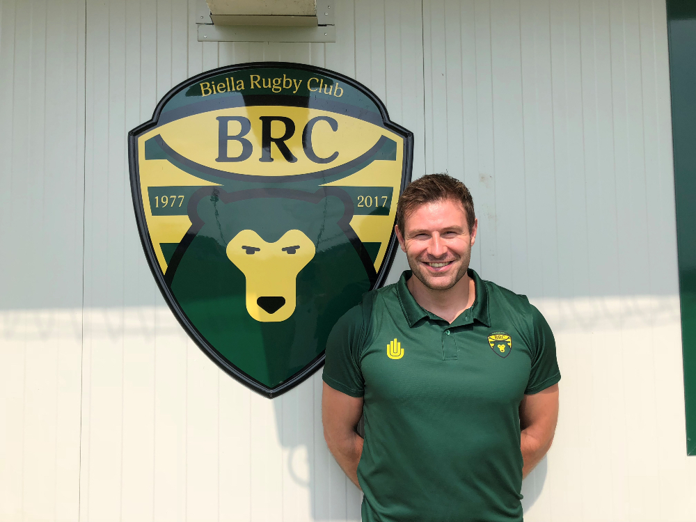 Fraser Murray next to the green and yellow Biella Rugby Club logo wearing a team shirt