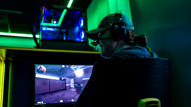 A games student wearing a cap sitting in front of a gaming PC with green lighting