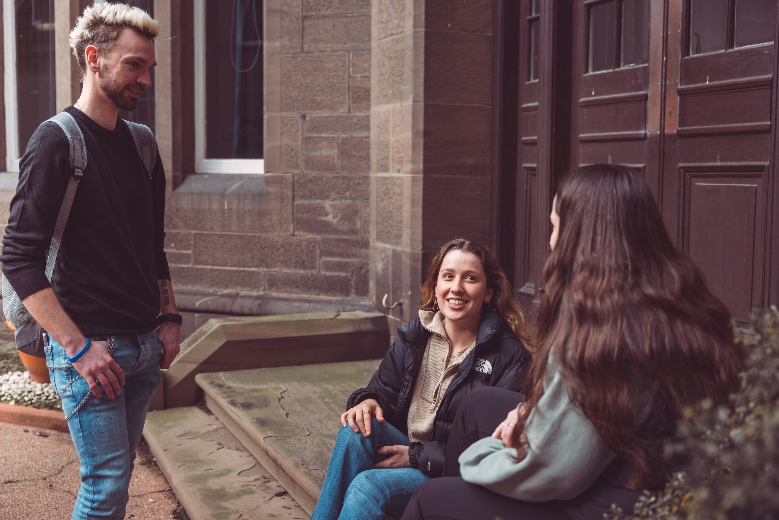 Three Students talking outside, two sitting on steps