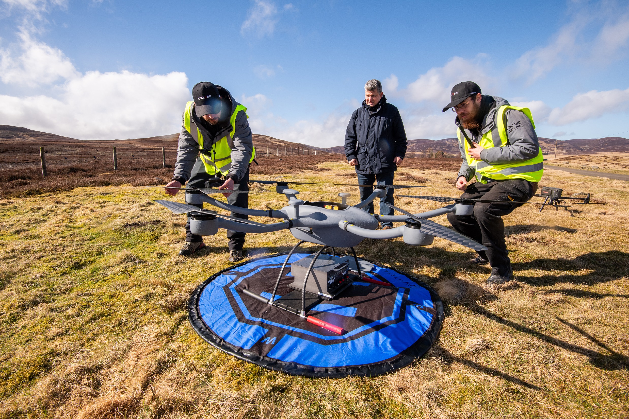 Two men in hi-vis vests and a man in a coat standing on a hillside looking at a large drone